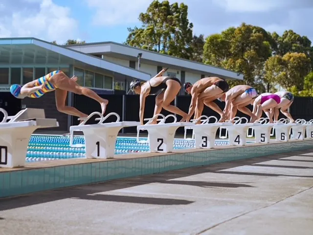 Six professional swimmers on starting blocks around an outdoor swimming pool jumping in to start a race.