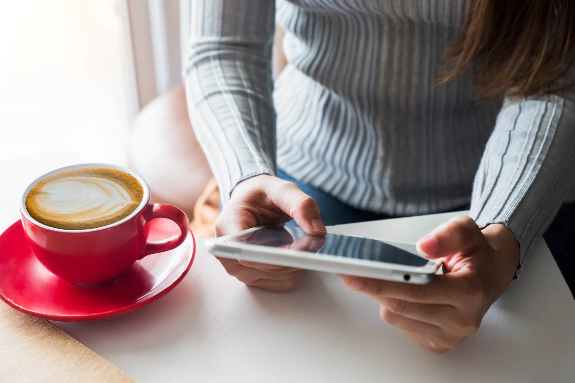 A cappuccino in a red cup sits on a table next to a women reading an e-magazine on her iPad.