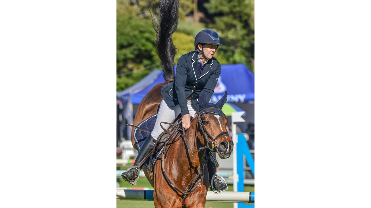 A lady dressed in the full helmet, jacket and jodhpurs on her horse and leaning forward as they horse has just jumped over a competition barricade.