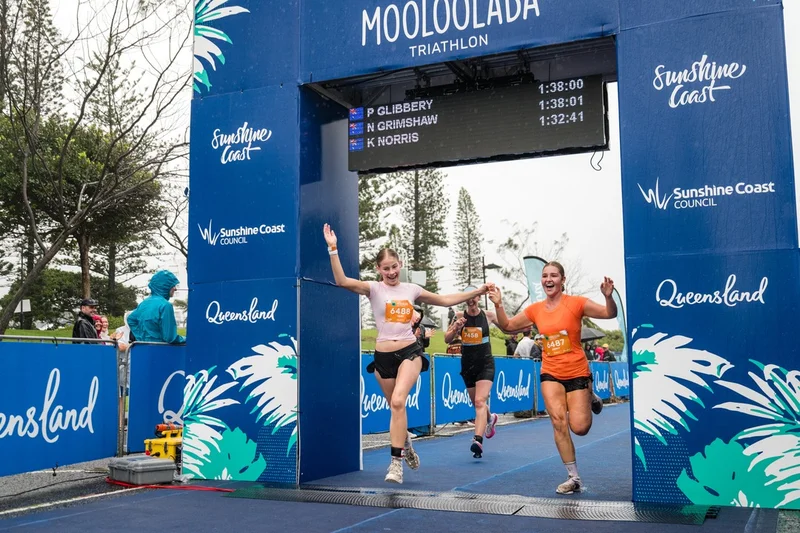 Mooloolaba triathalon finishg line. Three female runners one in white logo shirt and one in bright orange shirt coming through the finish line holding hands. Blue archway with logos Sunshine Coast COuncil and Sunshine Coast and Queensland.