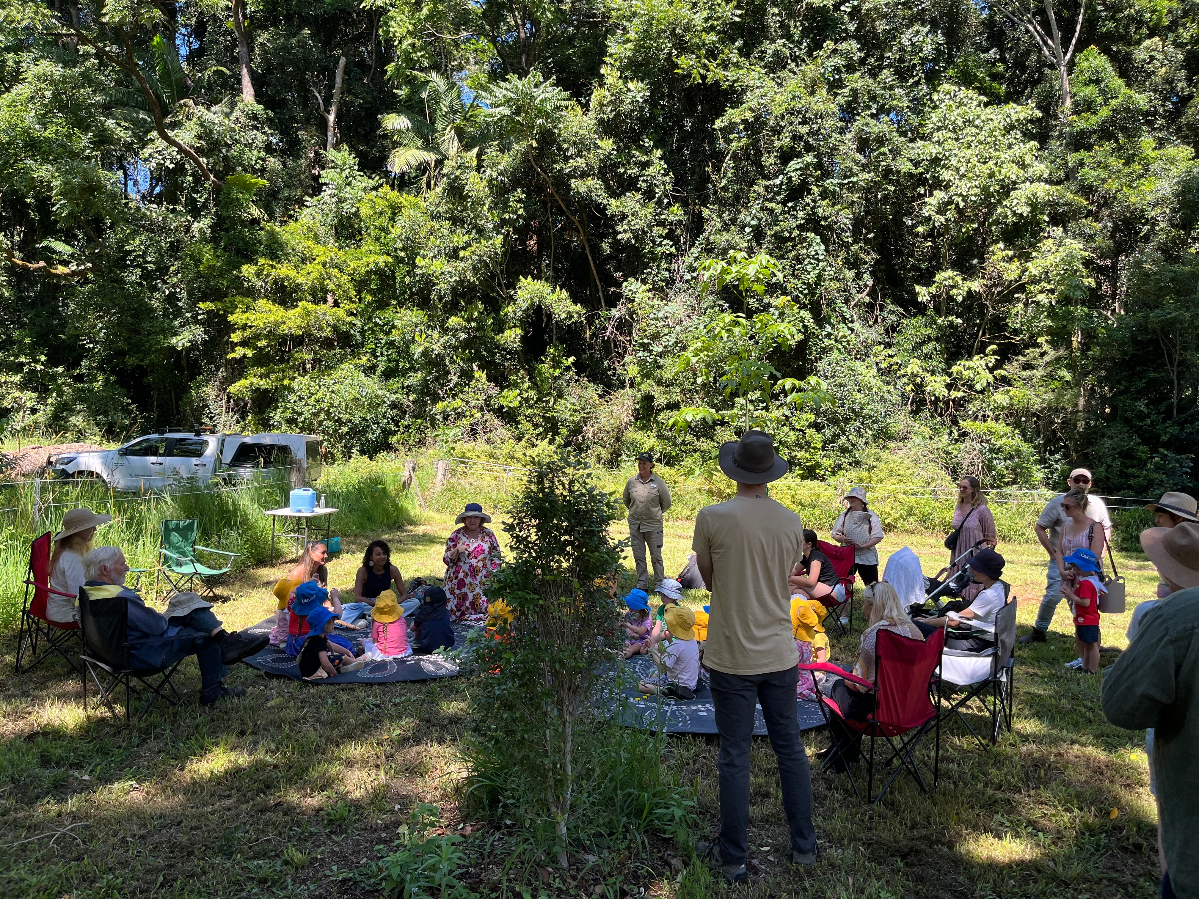 A crowd of people enjoy morning tea on picnic blankets in a grassy area with tall trees behind them.