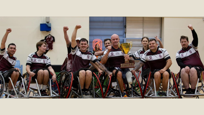Nine wheelchair rugby players (Qld team), some with an arm in the air, one man near the centre holding a gold trophy