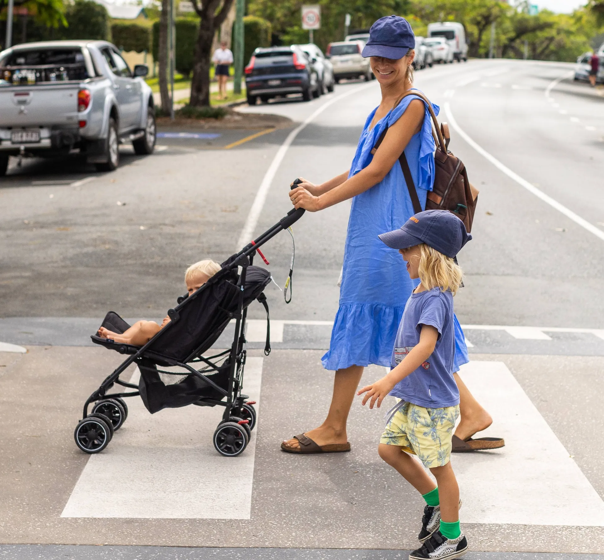 A little boy walks across a zebra crossing with his Mum who is pushing a troller with a baby.