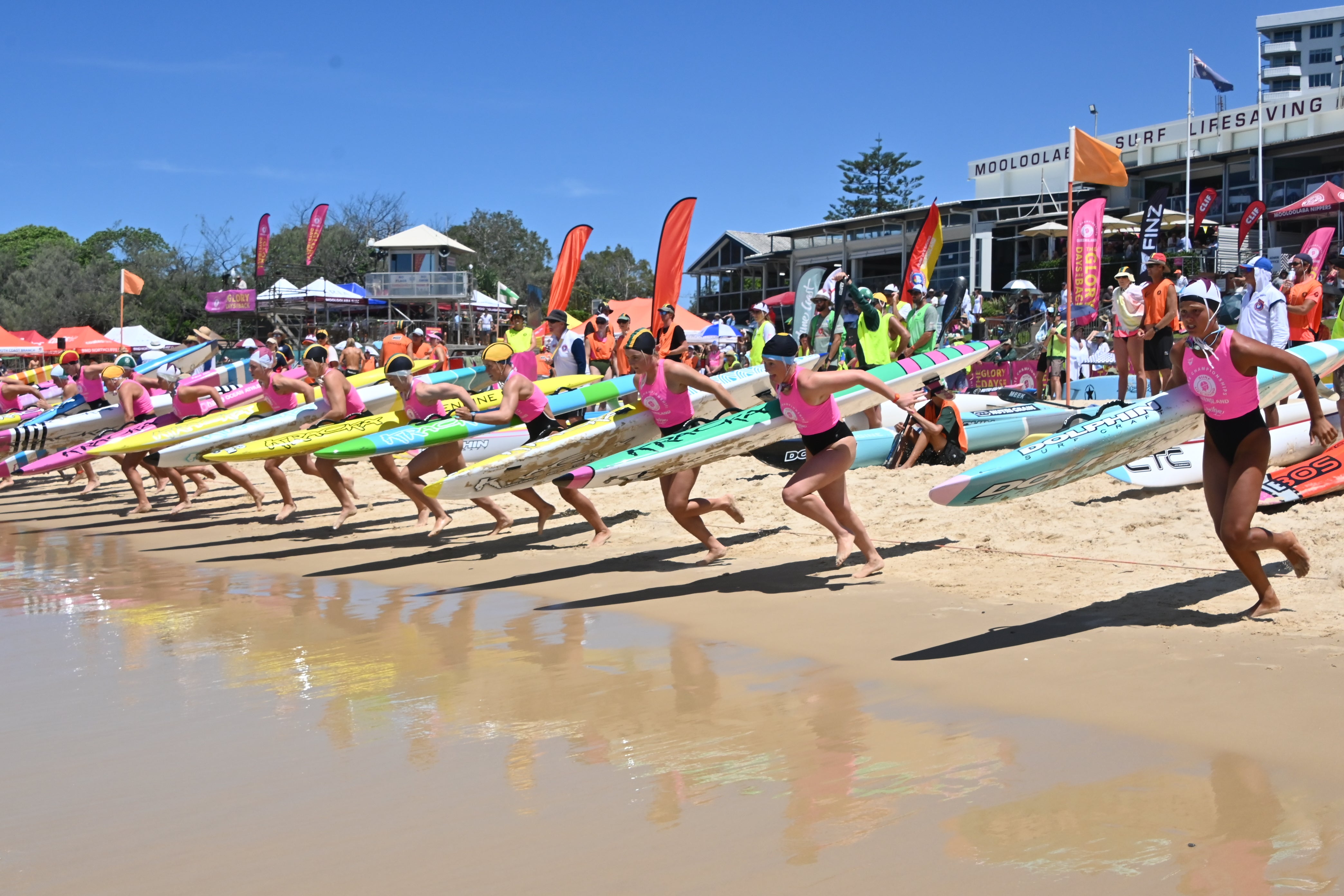 Surf Life Savers lined up with their boards ready to race into the water. Mooloolaba Surf Life Saving Club in the background.