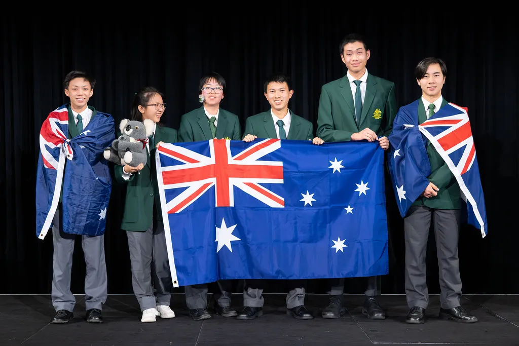 Students holding the Australian Flag, and two students on each end have the Australian flag wrapped around them. The 2nd student from the left is also holding a toy Koala. They are dressed in dark green blazers with grey plants.