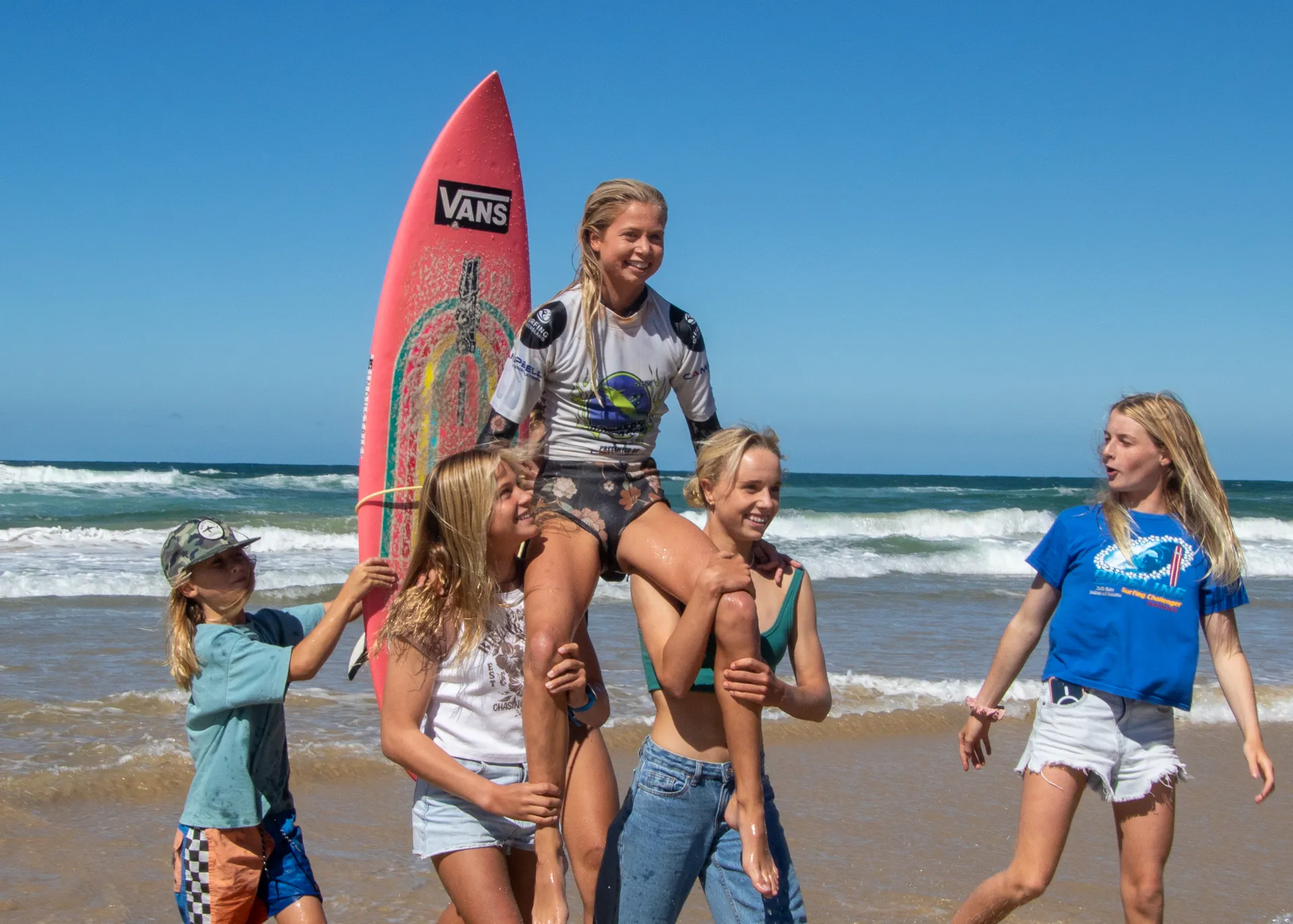 Surfing Qld Parko junior champion behing held up by fellow surf mates, with her surfboard behind her and the ocean behind them all.