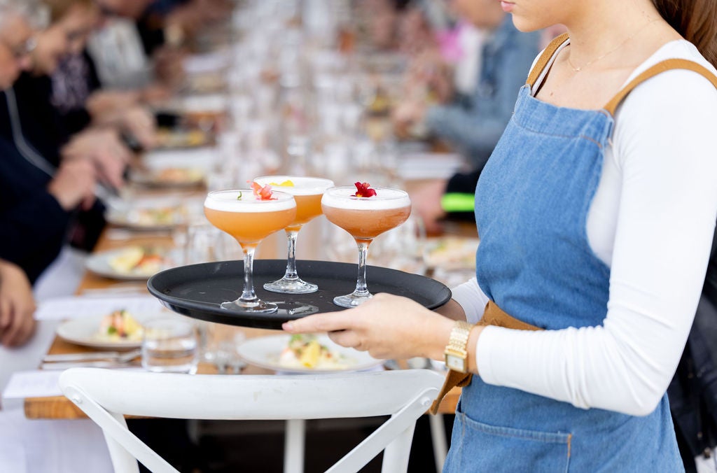 Three cocktails being served - blurred behind the try and server is a long table with guests eating food