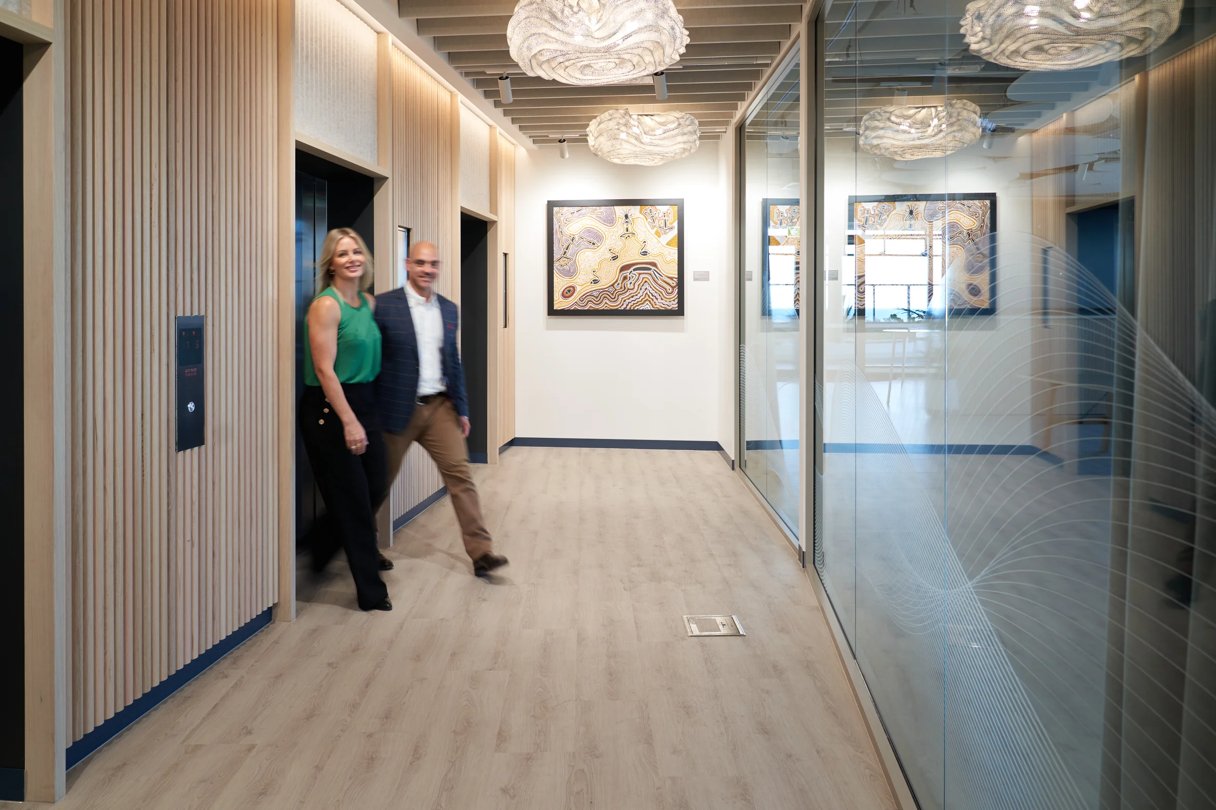 Professional couple, male and female arriving at Altitude Nine via the lift lobby. They are walking towards the camera, smiling with a First Nations artwork piece on the lobby wall behind them.