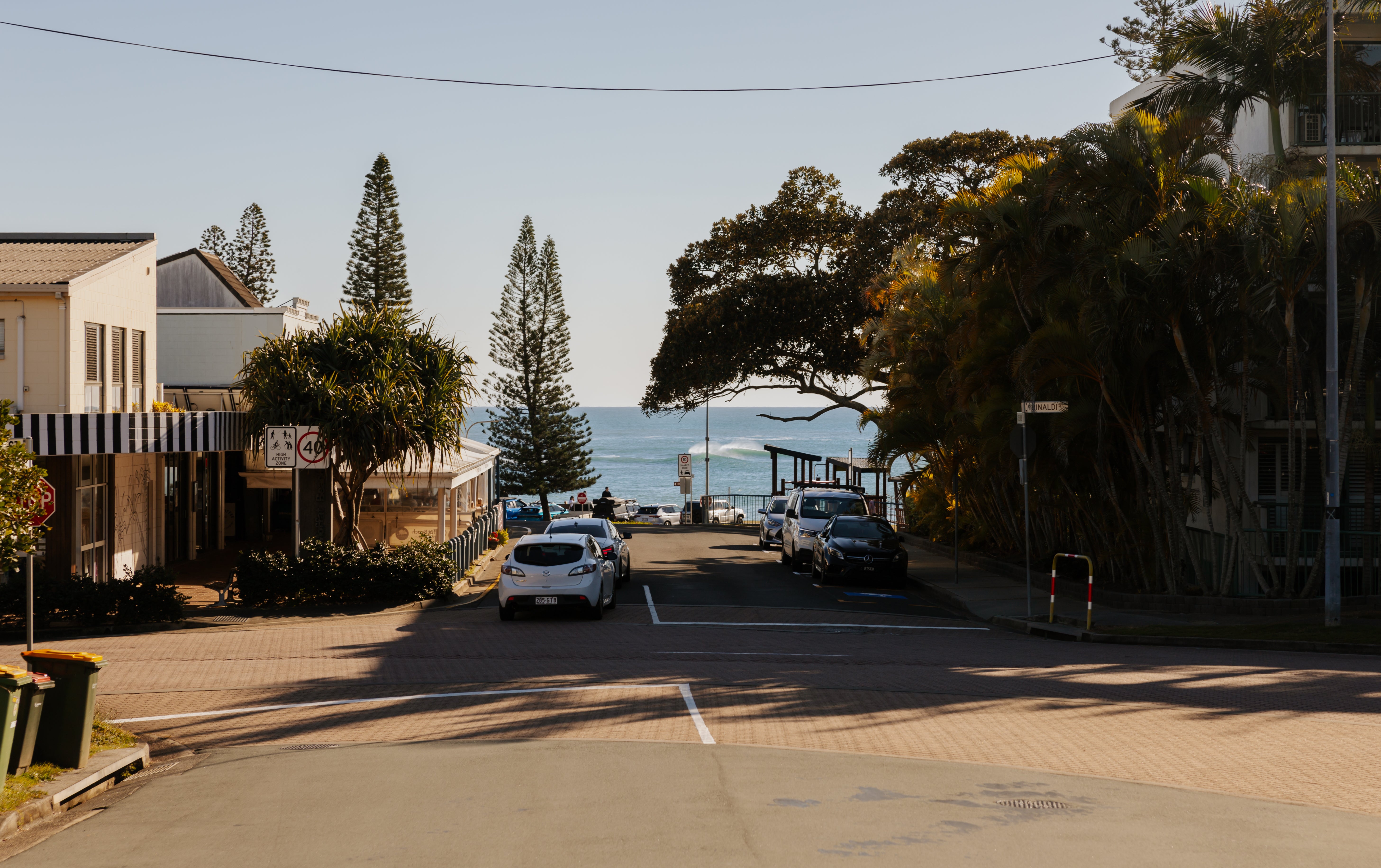 A bustling town centre featuring a road lined with cafes, shops, and trees. Cars are parked along the street, and in the background, a beautiful beach and ocean are visible.