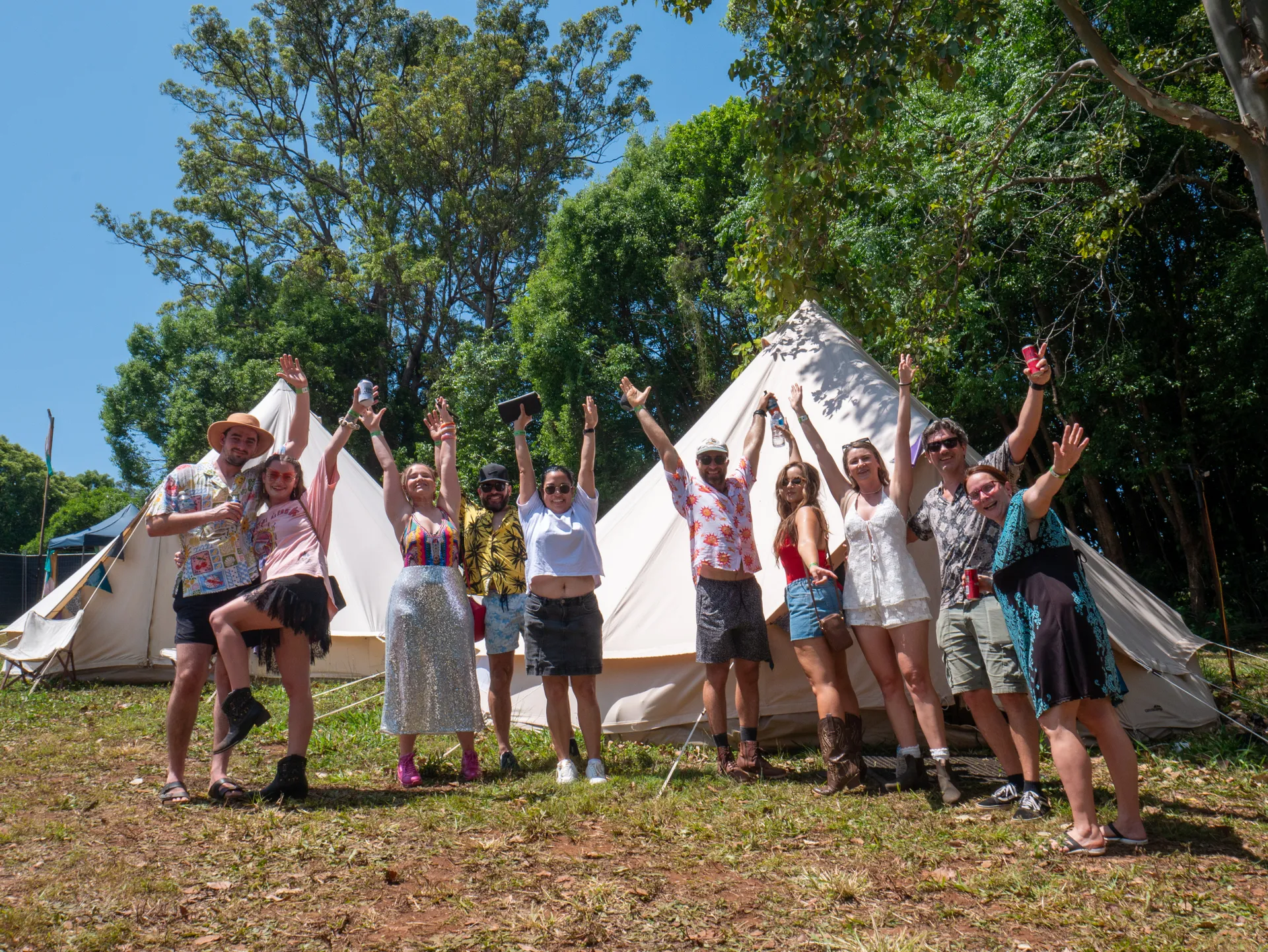 10 festival campers throwing their arms up in the air in front of two white tents at the festival campsite.