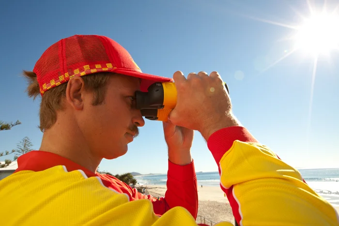 A man in the Surf Life Saving uniform long sleeve yellow shirt, red hat - holding binoculars up to his eyes. Blue ski background with the sunshine sparkling top right corner.