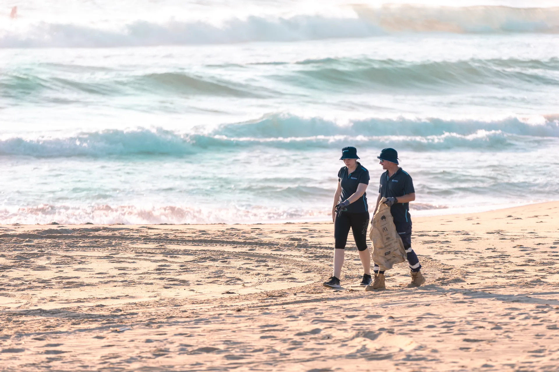 A group of people collecting rubbish on a sandy beach, working together to clean up the environment. They have a white bag in their hand to contain the rubbish.