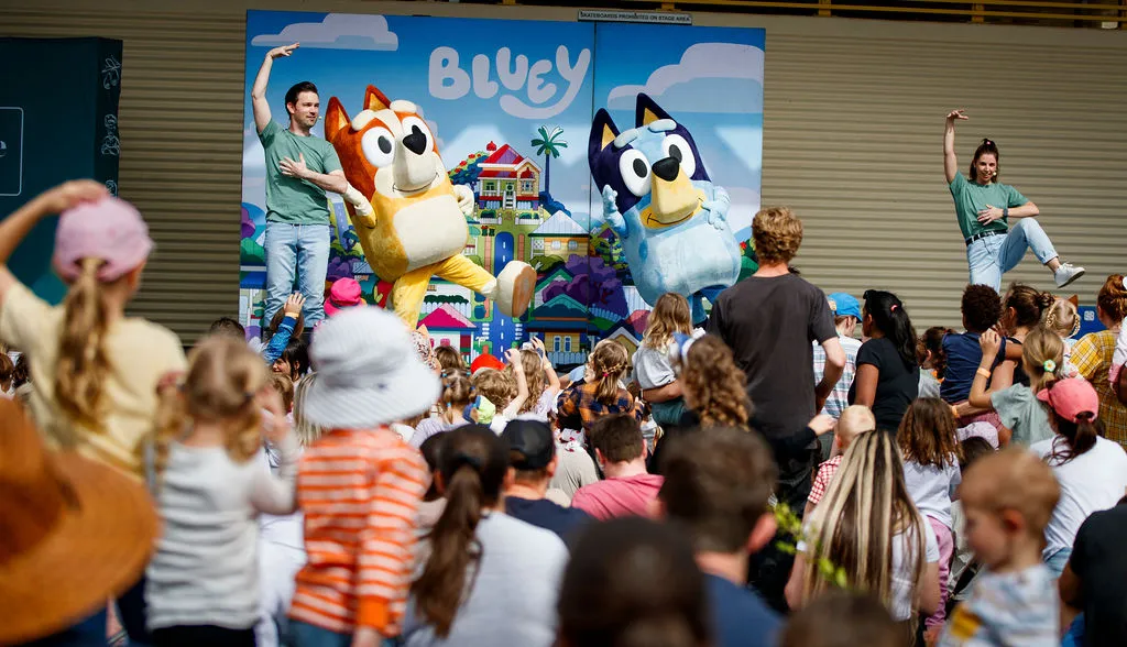 Kids Characters Bingo and Bluey on stage with lots of heads (the crowd) in the foreground of the image.