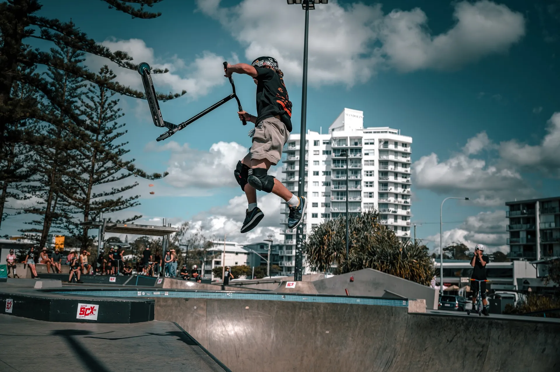 A guy executing a move with his scooter flipping around in the air, above a skatebowl. There is a tall white resort building in the background with blue sky and clouds.