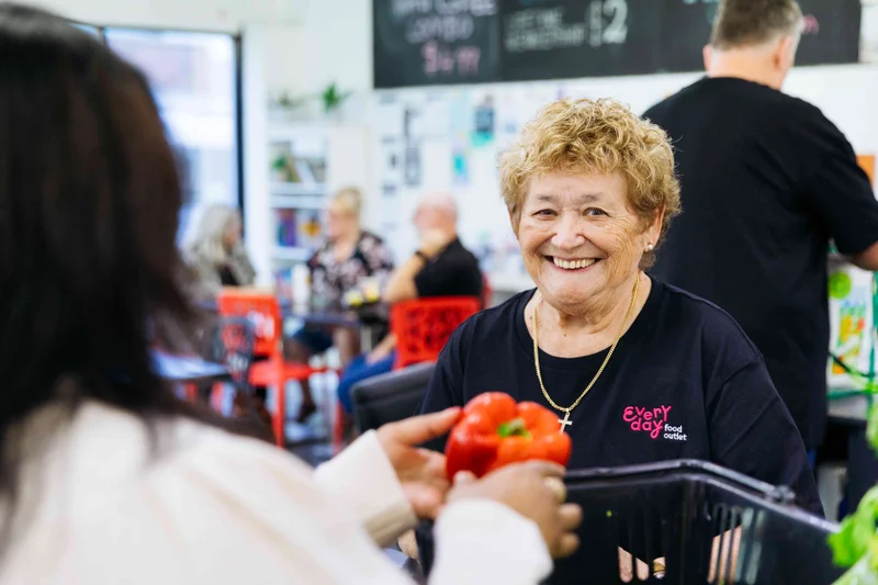 An older woman wearing a black shirt with a logo, smiles at the camera. The woman in front of her is washing a capsicum. 
There is a group of people also wearing black shirts, in the distance talking.