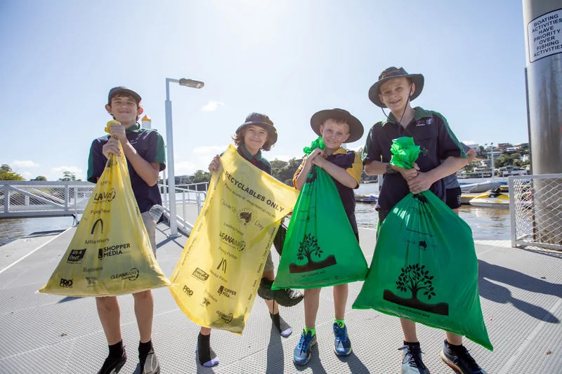 Four children looking at the camera smiling. Two on the left have yellow plastic bags in their hands, two on the right have green bags in their hands. the bags are full or rubbish. A river is in the background.