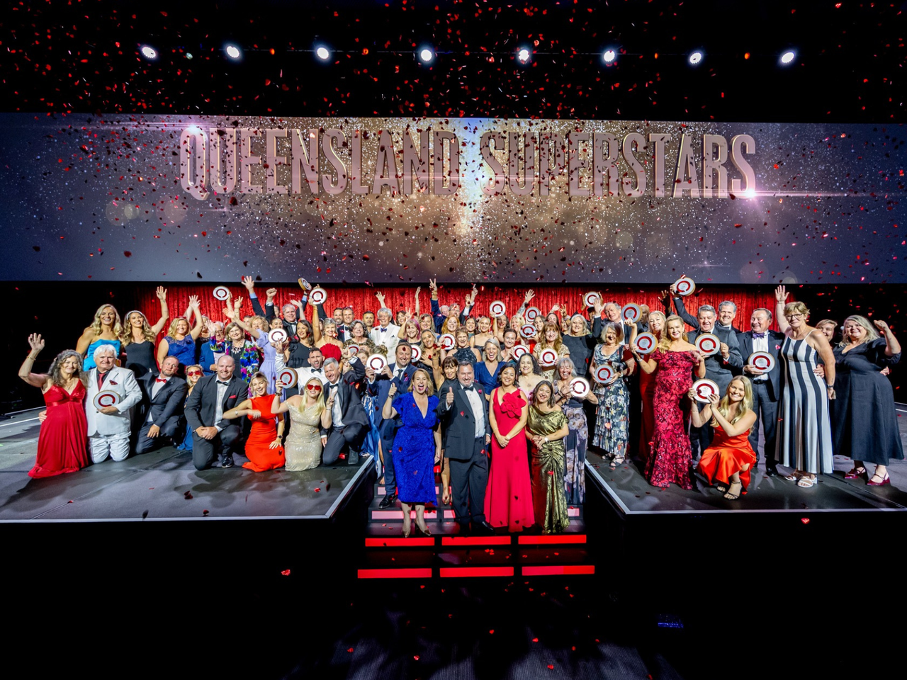 A group photo of all the winners on stage, with Queensland Superstars as the backdrop and confetti / glitter falling from above - making everything look a little blury.