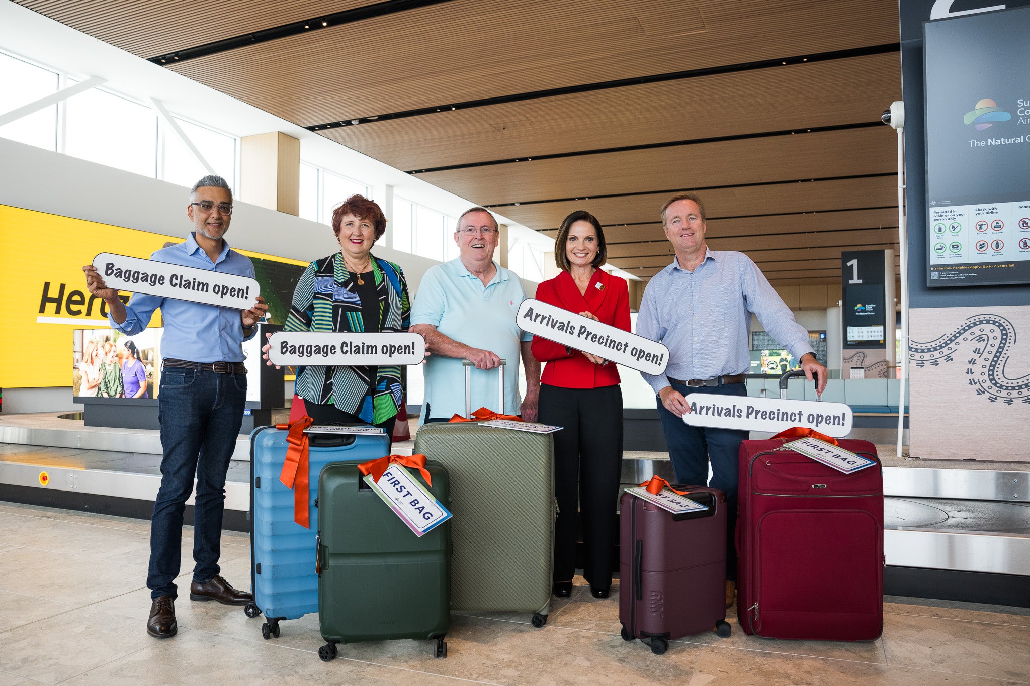 People holding signage (Baggage Claim open! / First Bag / Arrivals Precinct open!) and standing in front of suitcases