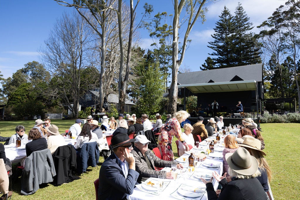 In an outdoor setting, this photo captures two long tables filled with guests, and a stage in the background