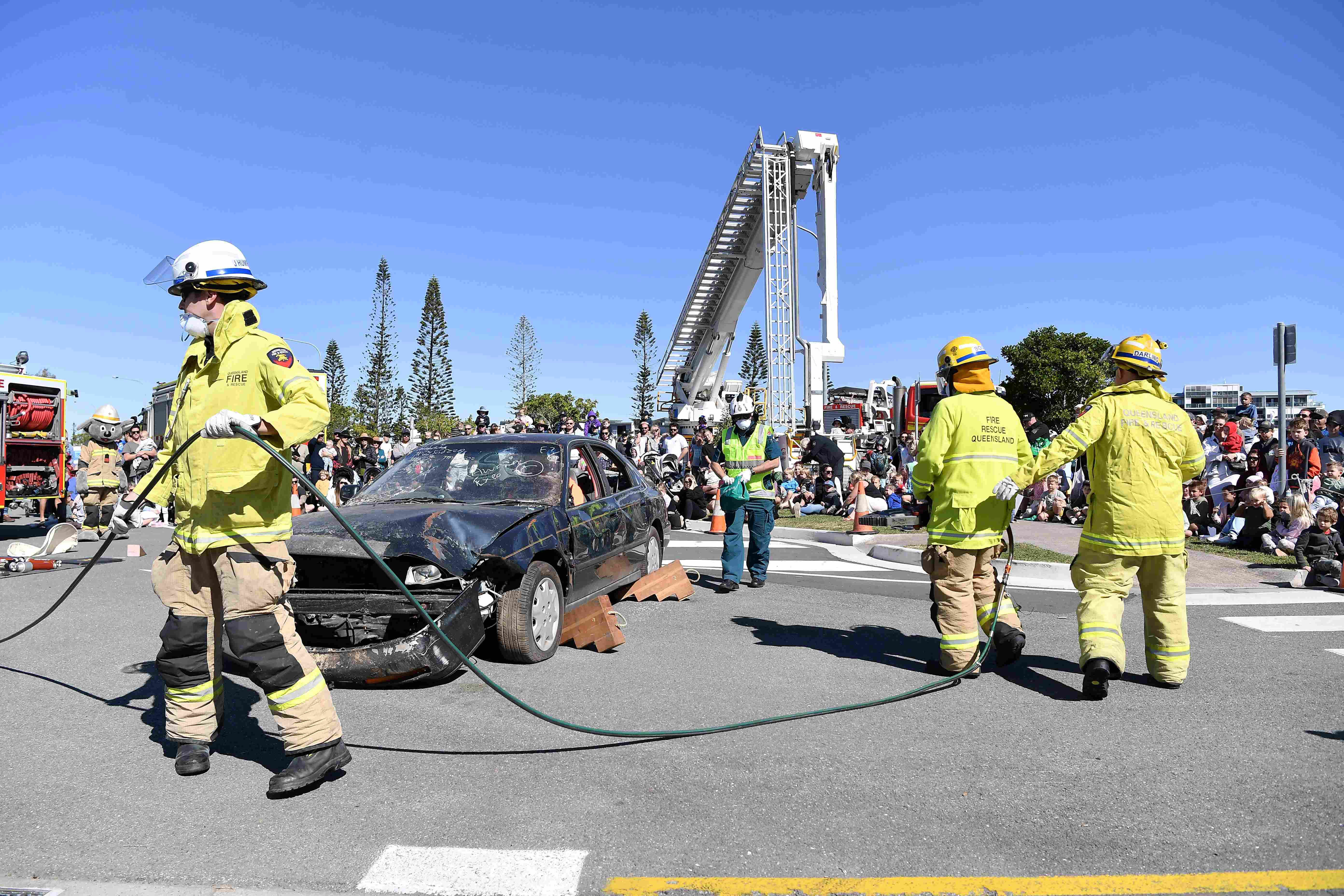 Four firefighters holding equipment surround a smashed car. A large crowd of onlookers encircle the demonstration.