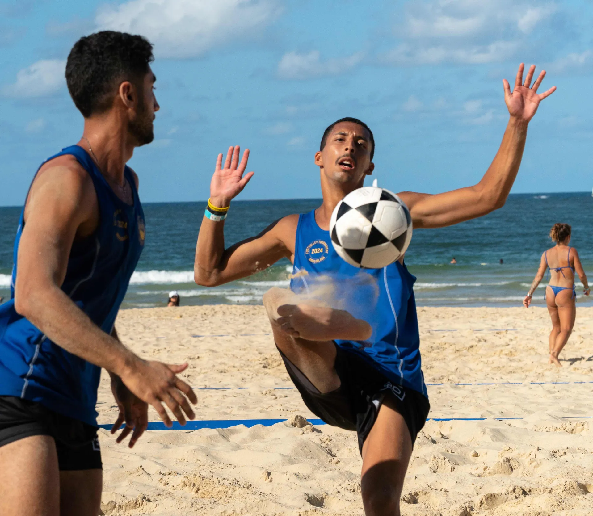 Two men wearing blue singlets are on the beach beside a volleyball net. The man on the right holds his hands up and kicks the ball with his foot.