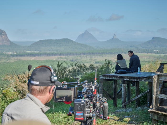 Jana McKinnon (Zoe Jacobs), Vinnie Bennett (Joseph Lim), cameraman at mountain lookout. Looking out to lush green feield and three mountains with blue skies and a few white clouds.