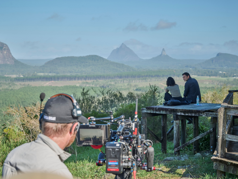 Jana McKinnon (Zoe Jacobs), Vinnie Bennett (Joseph Lim), cameraman at mountain lookout. Looking out to lush green feield and three mountains with blue skies and a few white clouds.