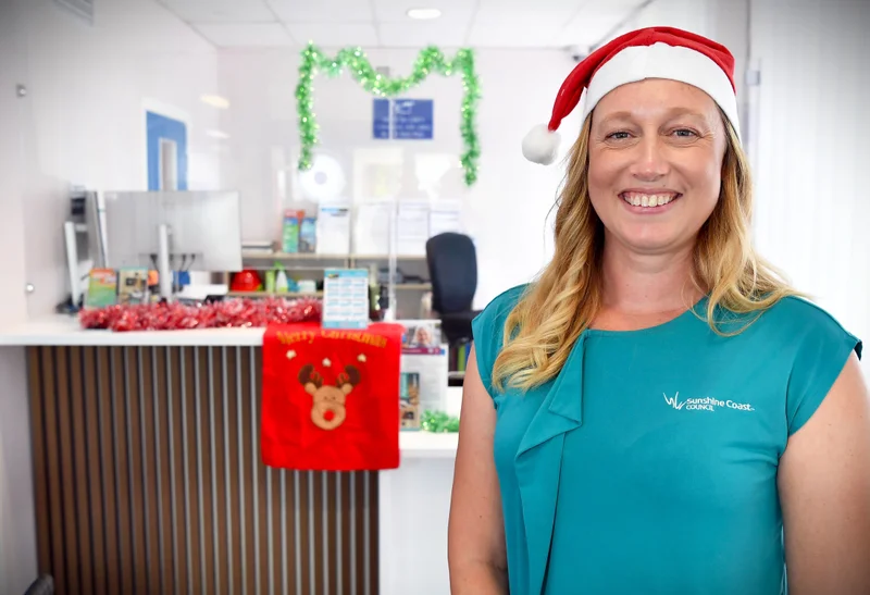 A friendly, smiling customer service Council worker wearing a Santa hat, stands in front of a customer service counter that's been decorated with Christmas tinsel.