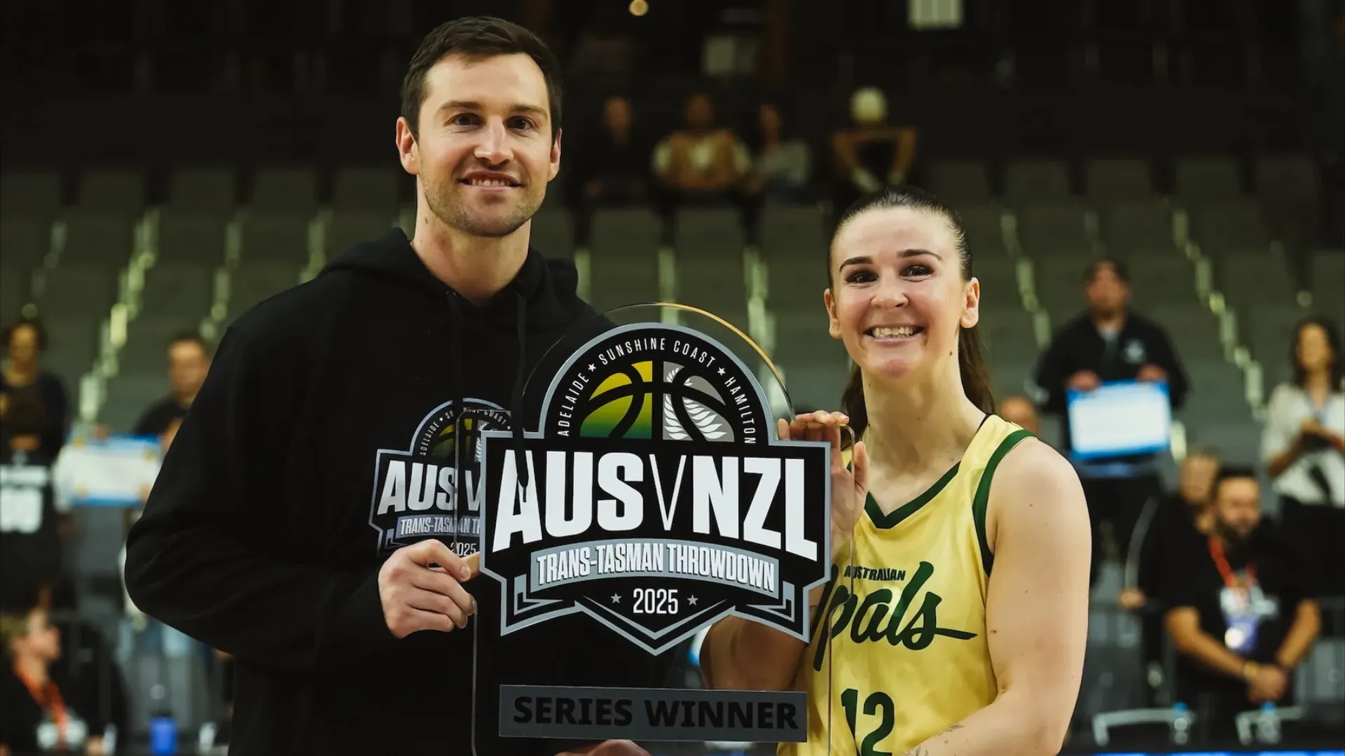 Two people (Australian Boomers and Opals players) holding the AUZ v NZL Trans-Tasman Throwdown 2025 trophy with blurred vision of stadium seating behind them.