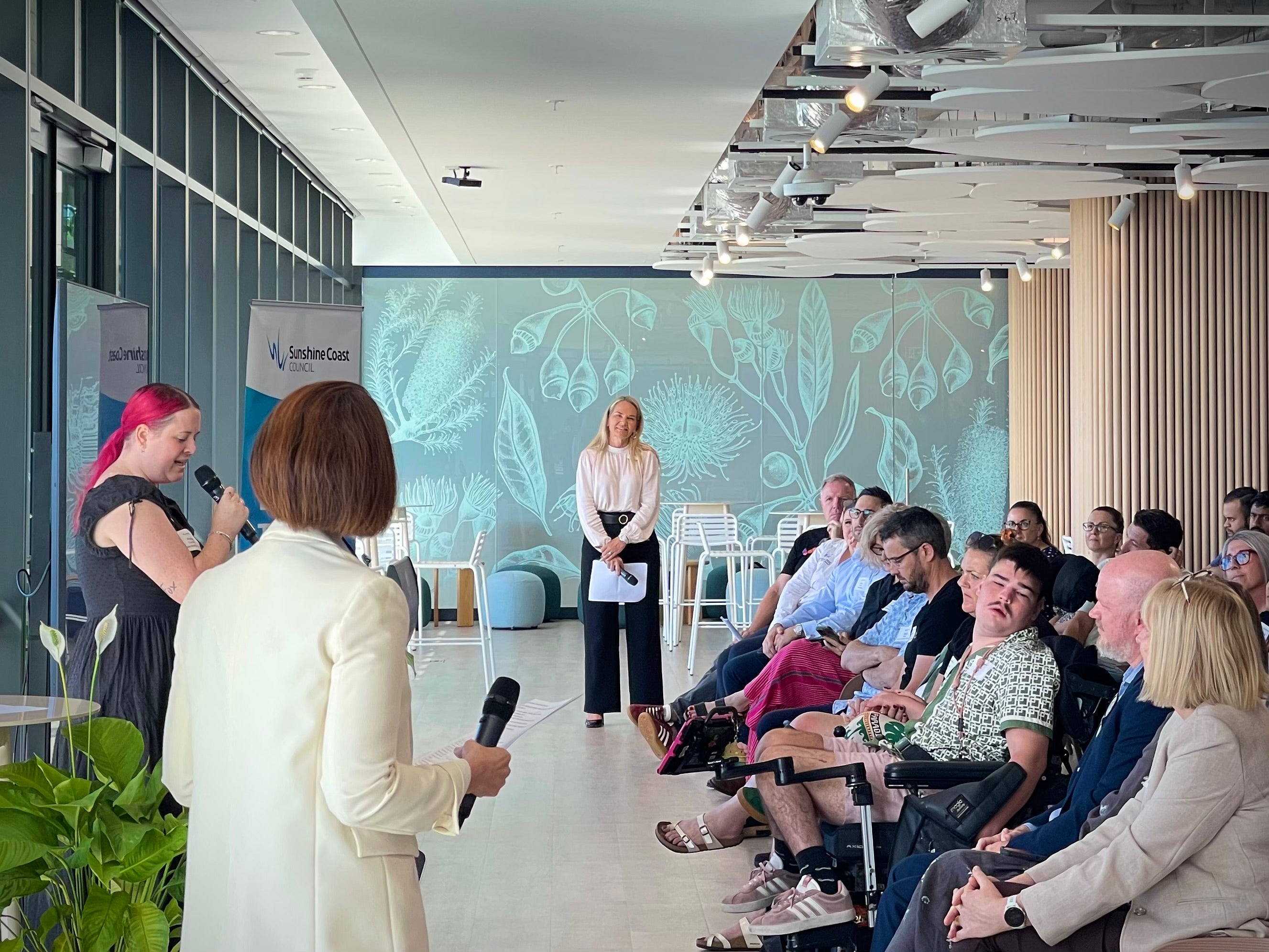 A woman with pink hair stands behind a podium talking to a room full of people seated in front of her. Two other women are standing and listening.