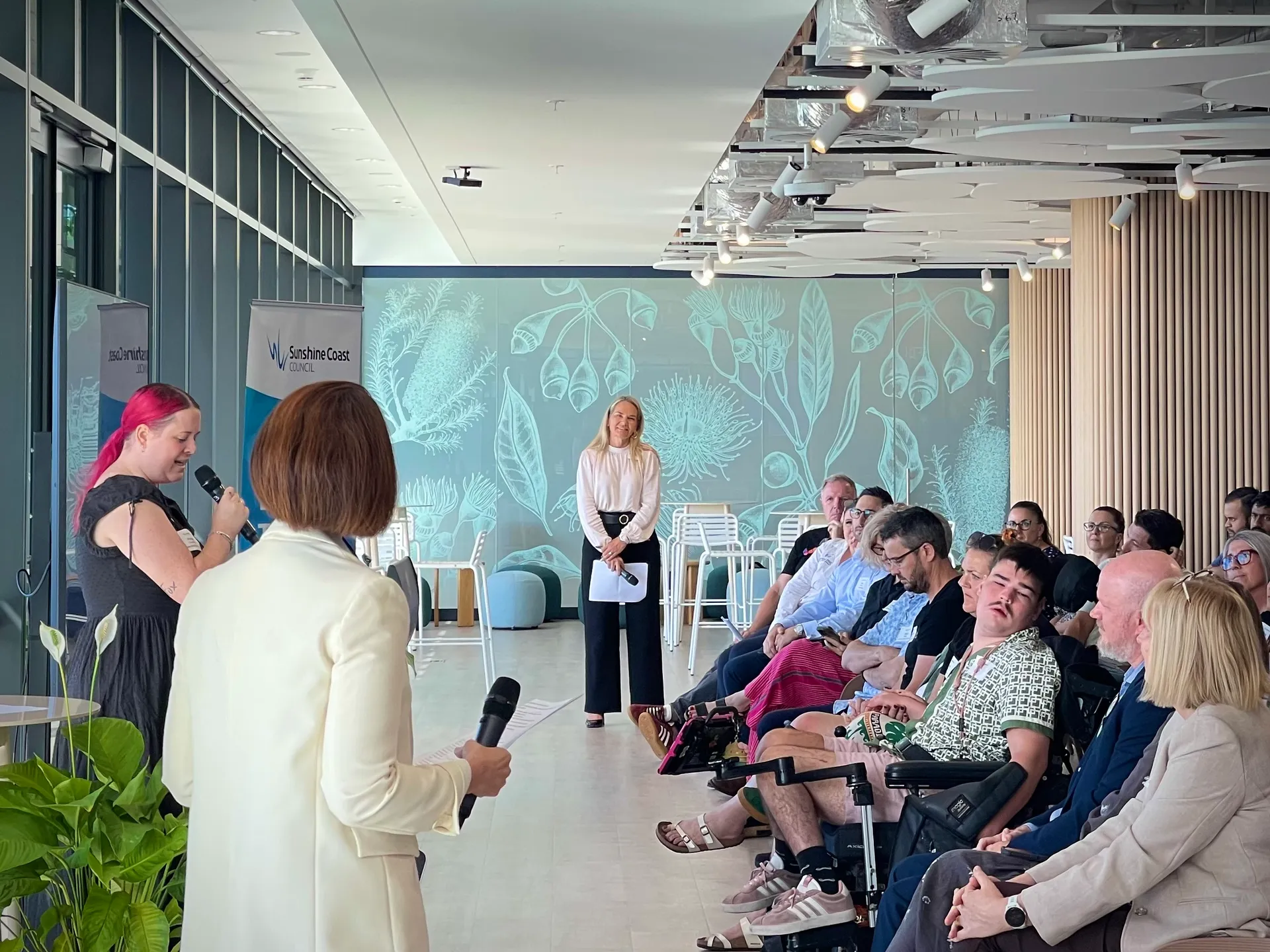 A woman with pink hair stands behind a podium talking to a room full of people seated in front of her. Two other women are standing and listening.