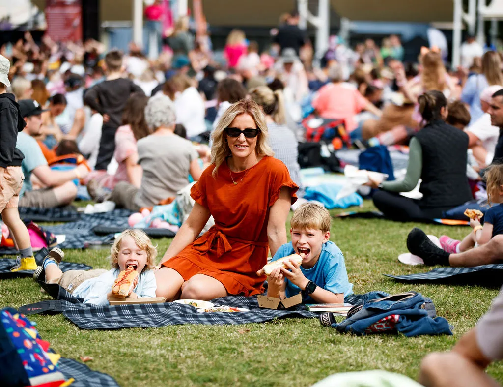 A mum on a picnic blanket with her two chilren, who are holding and about to bite into a hotdog.