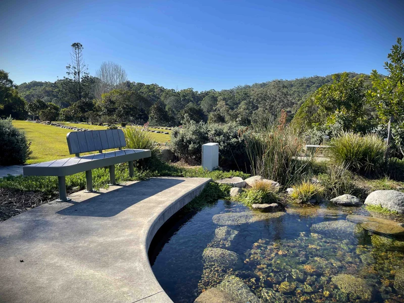 Beside a pretty pond surrounded by plants, is an empty garden chair. IN the background are gravestones, rolling green hills and tall trees.