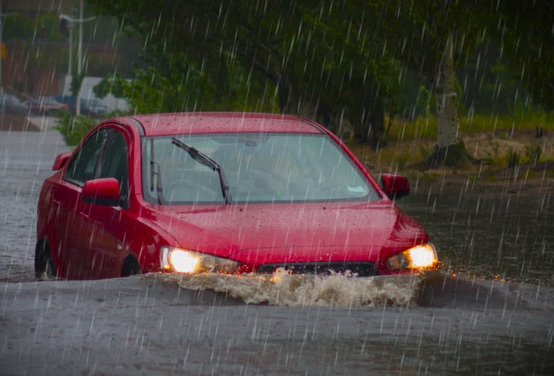 A red car is immersed in flood water that is covering a suburban road.