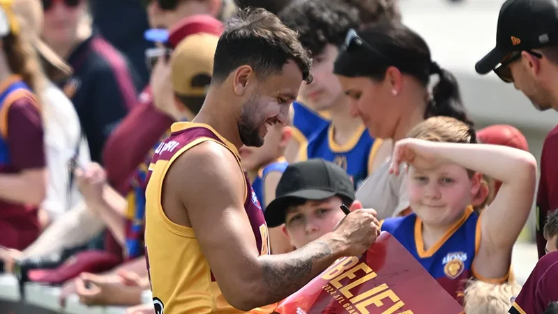 A Brisbane Lions player signing fan merchandise, with crowd in the background.