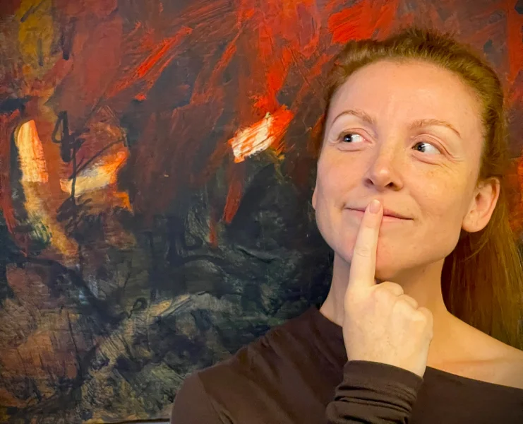 A young, brown eyed, red headed woman stands in front of a red and brown abstract painting with a pointer finger on her mouth and looking cheekily to the side.