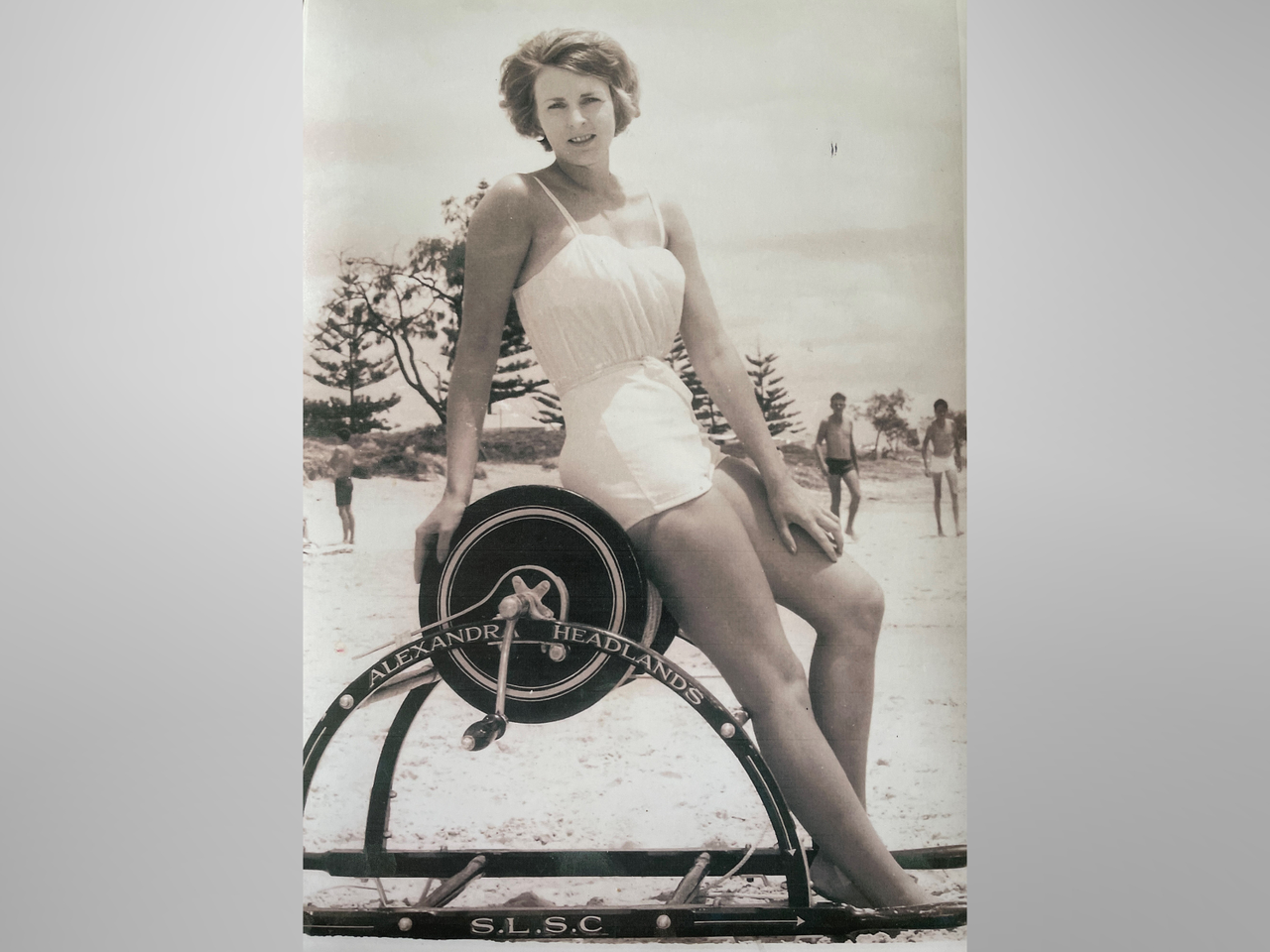 1960s sepia photo of a woman in a white one-piece swimming costume posing barefoot on a sign for Alexandra Headlands S.L.S.C.