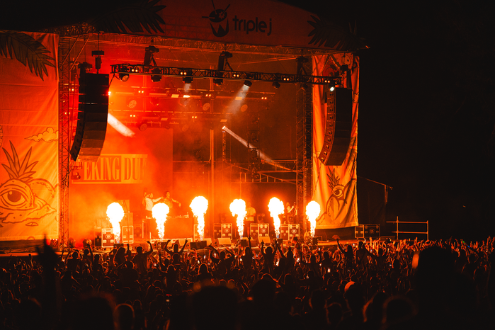 A stage bathed in a red glow, with two speak stacks hanging from the roof, and six fire balls coming up from the bottom of the stage, with a crowd in the dark in front of the stage.
