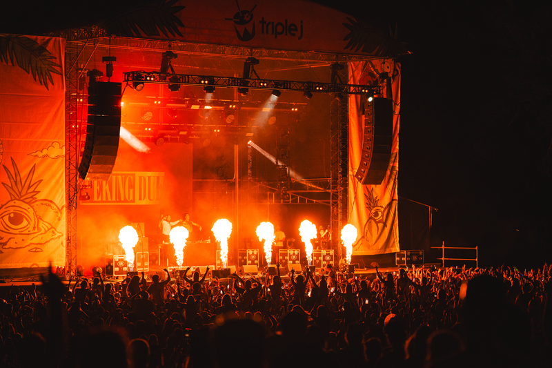 A stage bathed in a red glow, with two speak stacks hanging from the roof, and six fire balls coming up from the bottom of the stage, with a crowd in the dark in front of the stage.