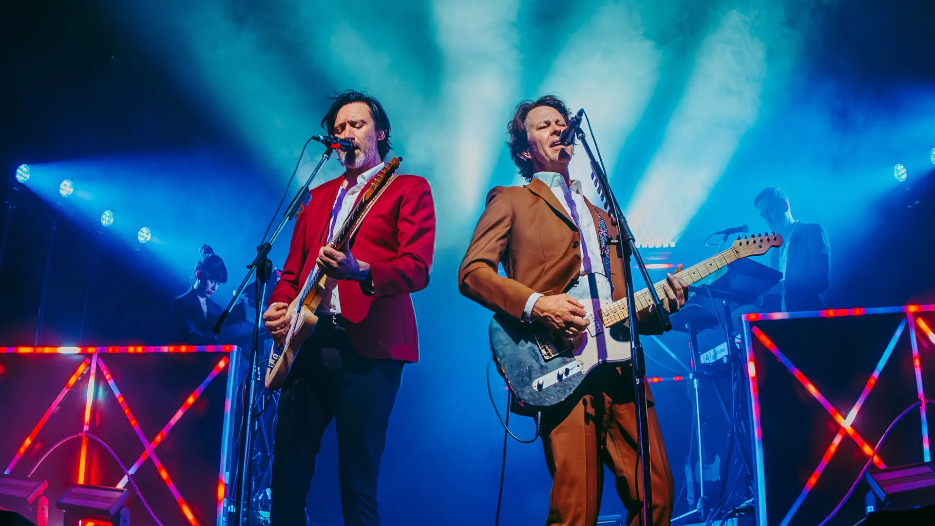 Bernard Fanning and Paul Dempsey playing electric guitars and singing. Blue lights behind them with moody music venue vibes.
