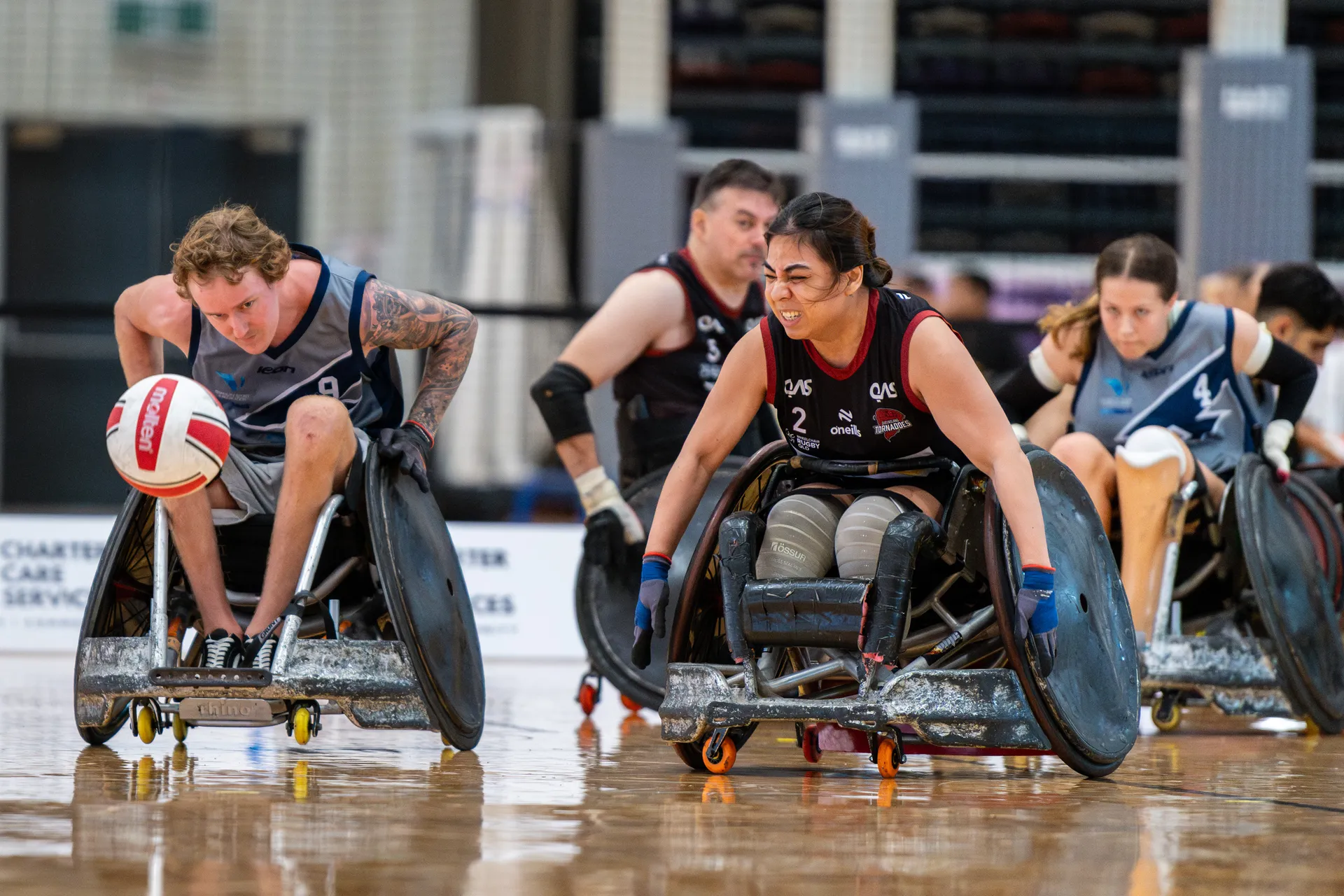 Multiple parasport athletes on the court, chasing a ball on the left.