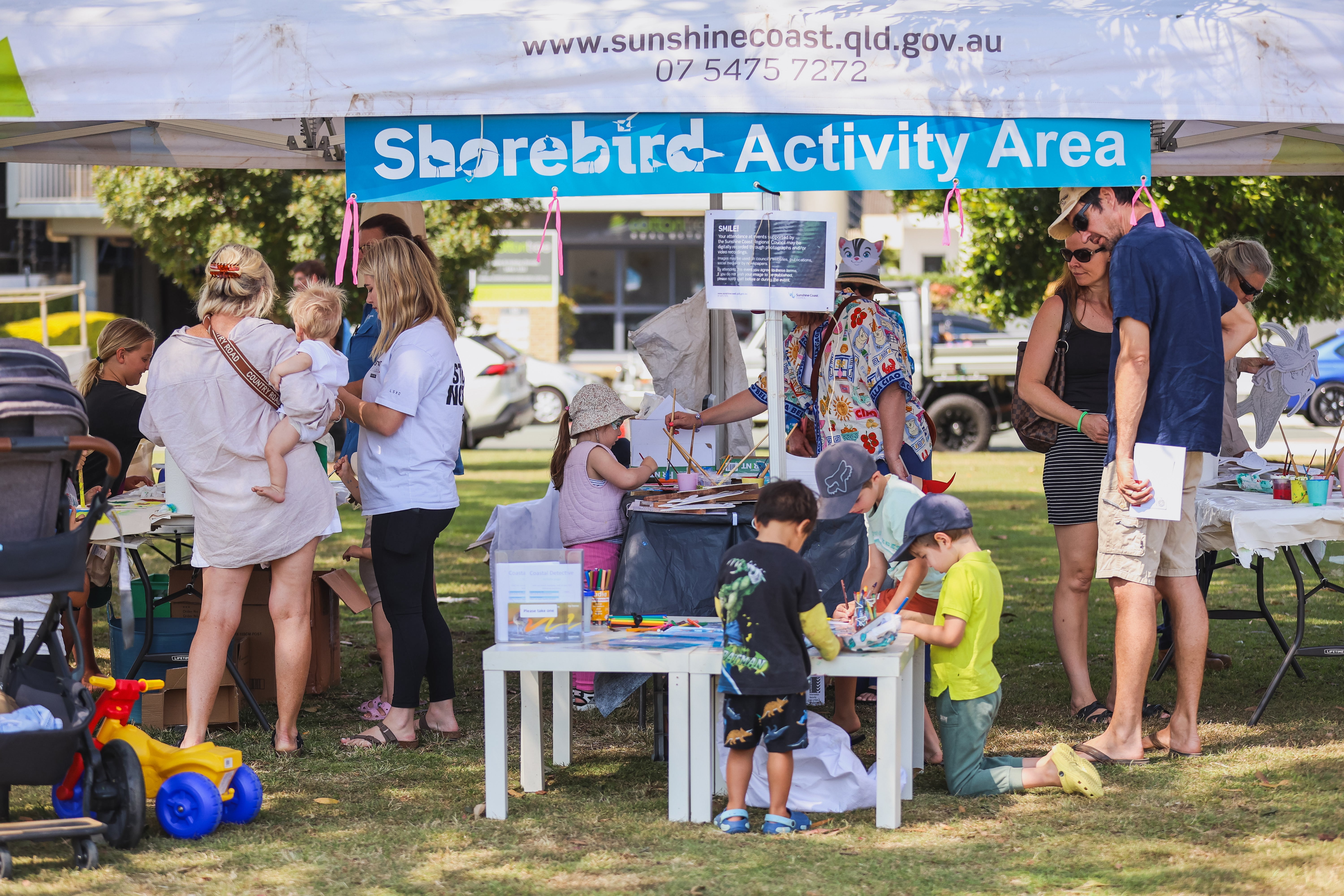 "People enjoying a shorebird festival on a sunny day in a park, with children painting and getting face paint, adults chatting and learning about migratory birds, and various activity stalls visible in the background."