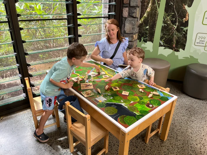 Two children sitting at low table playing a game with their female caregiver watching on. They are inside a large building with glass windows and vegetation outside.