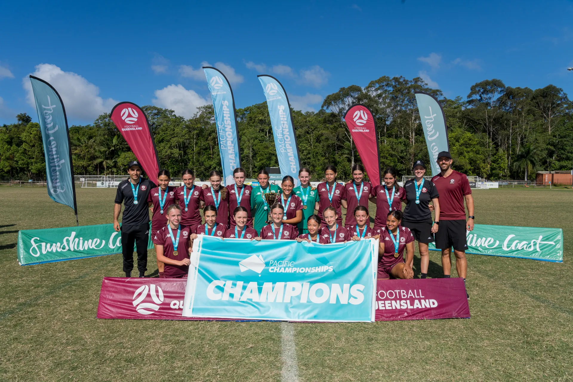 A group of junior players with medals around their neck plus referees, and coach. The front row are holding a sign that says Pacific Championships - CHAMPIONS, with a number of vertical flags in the background - Football Queensland, Pacific Championships, Sunshine Coast