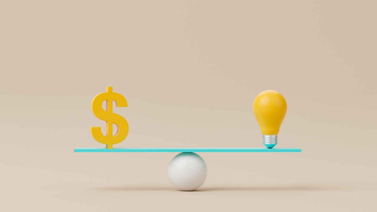 balance scale with a light bulb one end and a dollar sign the other