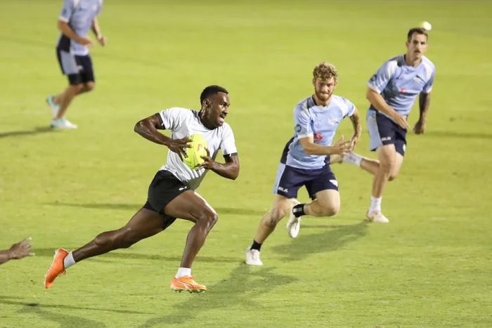 Rugby player running with a yellow rugby ball in a white shirt with two other players in blue shirts getting running towards him to tackle. On a lush green football field. 