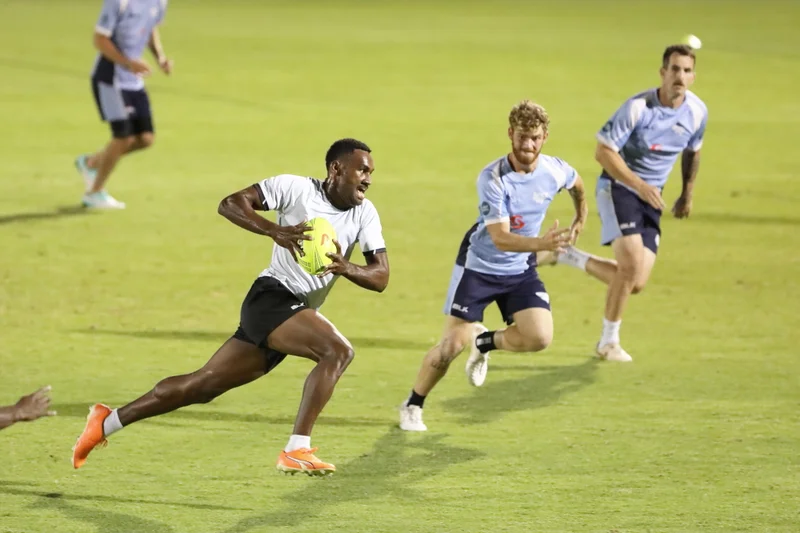 Rugby player running with a yellow rugby ball in a white shirt with two other players in blue shirts getting running towards him to tackle. On a lush green football field.