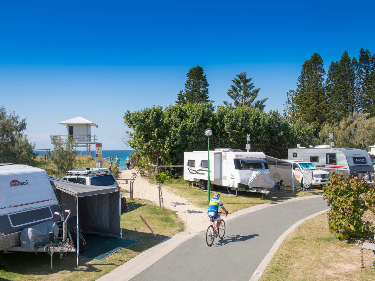 An image with caravans, campervans, tents and someone riding a bike on the main path through a holiday park, with a sandy pathway to the beach and a lifeguard tower