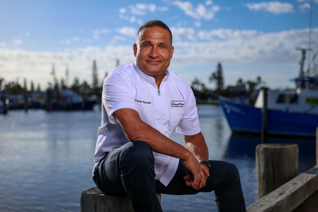 Chef Peter Kuruvita sitting wharf side with blue sky and clouds above, boats behind him and water in the background.