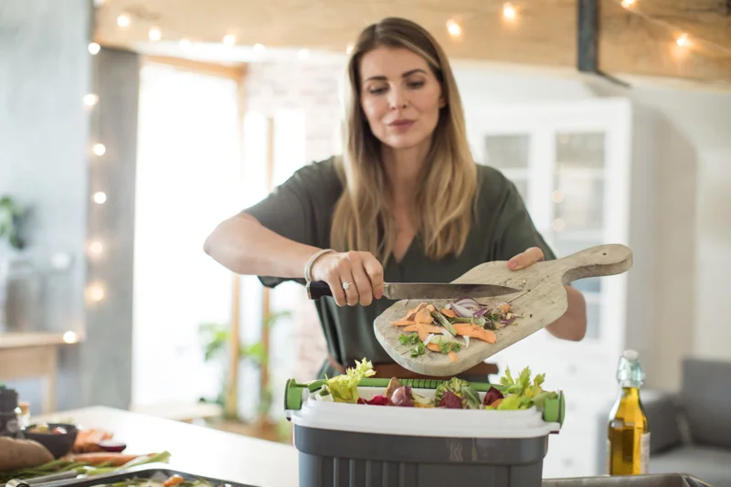 A lady in the kitchen is scraping scraps into a recycling container.